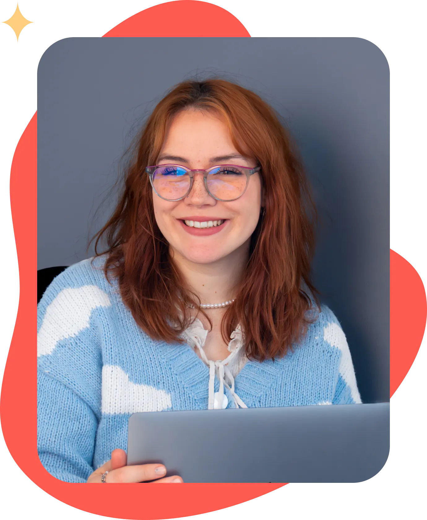 Smiling person with glasses and red hair, wearing a blue and white sweater, holding a laptop against a gray background with a red border.